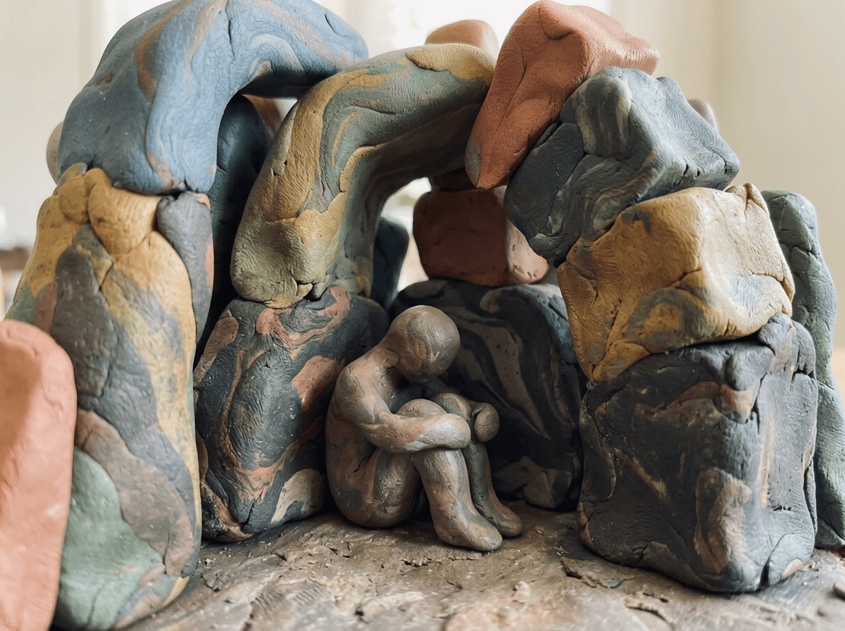 Small clay human figure sitting with arms around knees inside a cave-shaped arrangement of multicolored clay blocks