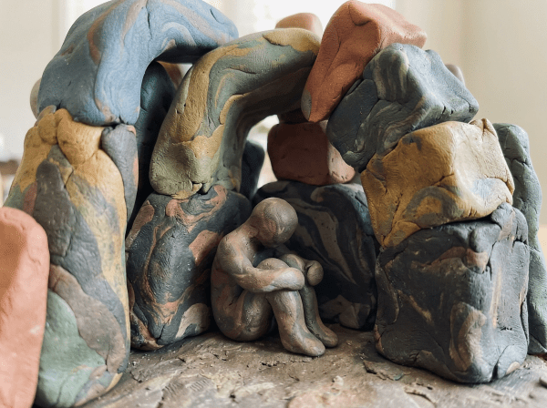 Small clay human figure sitting with arms around knees inside a cave-shaped arrangement of multicolored clay blocks