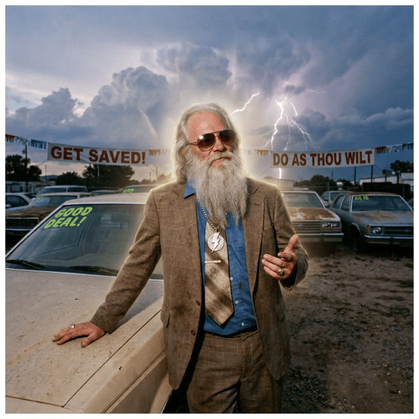 Man in suit at car lot beside cars labeled “GOOD DEAL!”, “BARGAIN CARS,” and “AS IS”